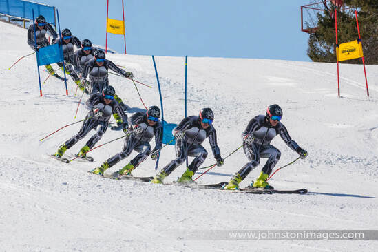 A total of eight photographs superimposed over each other to create one composite image of a ski racer skiing on the slopes. The image shows their progression as they race down the hill. It is a man in a ski racing suit that is black, white and grey. He has bright yellow ski boots, skis, poles, a helmet, and goggles on. He is skiing over white snow past gates, which are poles in the ground with flags on them, which defines the course. The sky is blue and there are a few pine trees off to the upper right of the hill.